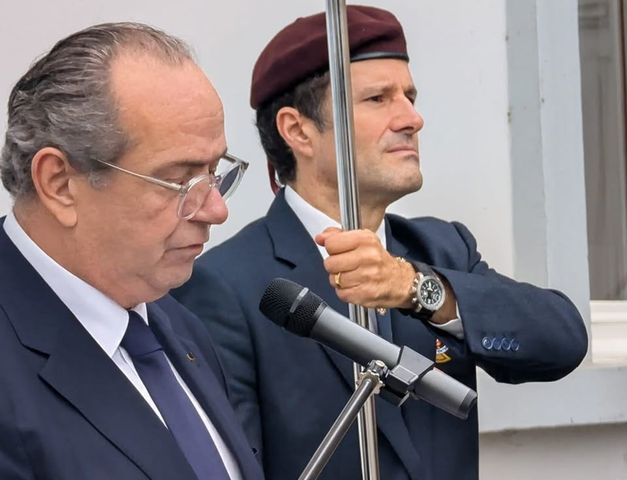 Dois homens em trajes formais participam numa cerimónia solene; um lê ao microfone enquanto o outro segura uma haste de bandeira, ambos com expressão séria, simbolizando homenagem e respeito aos soldados portugueses caídos na Primeira Guerra Mundial.