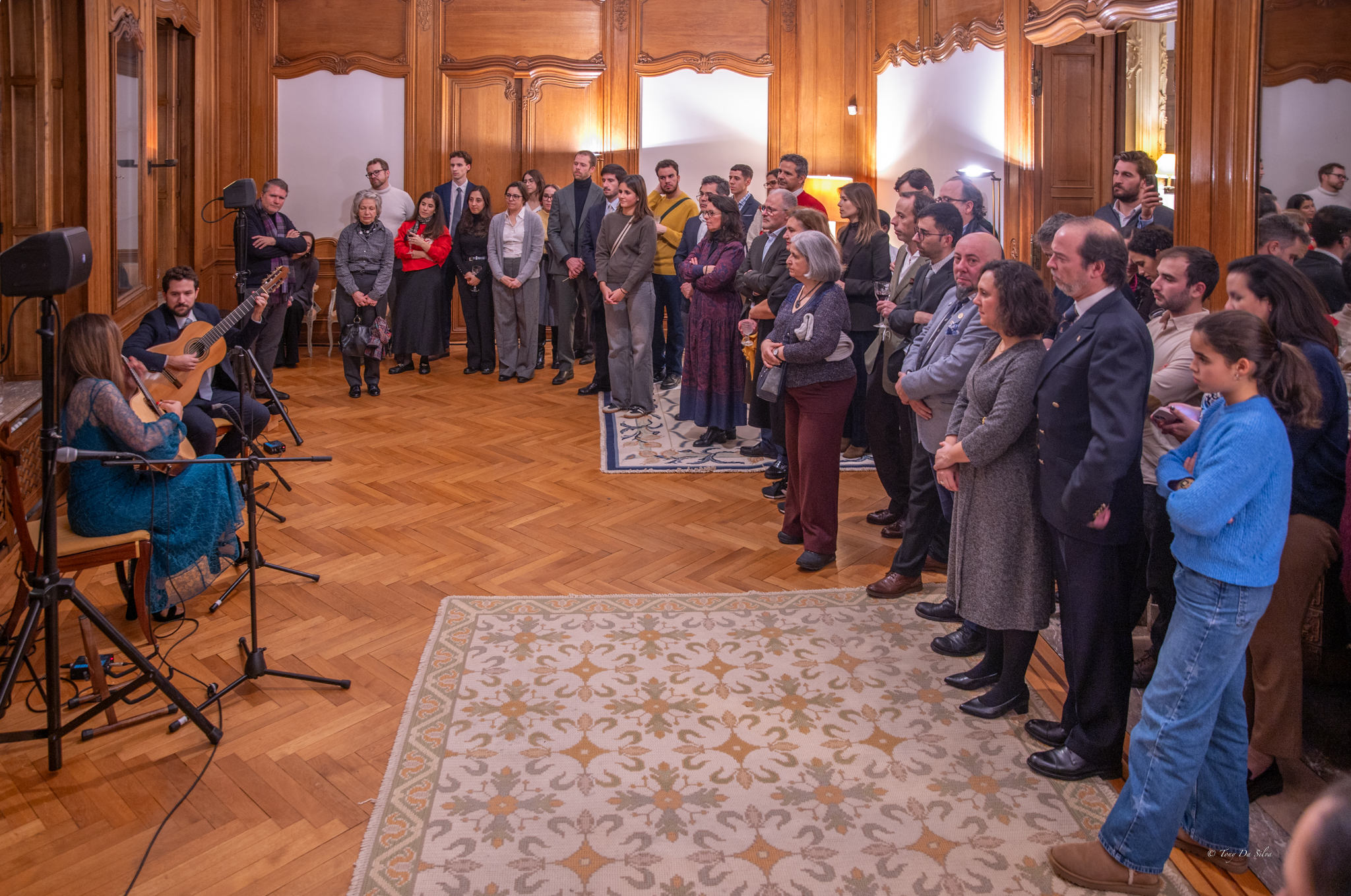 Concerto de Natal na residência oficial do Embaixador de Portugal na Bélgica, com Marta Pereira da Costa a tocar guitarra portuguesa perante uma plateia numerosa e atenta, num salão elegante, celebrando a cultura e comunidade portuguesa.
