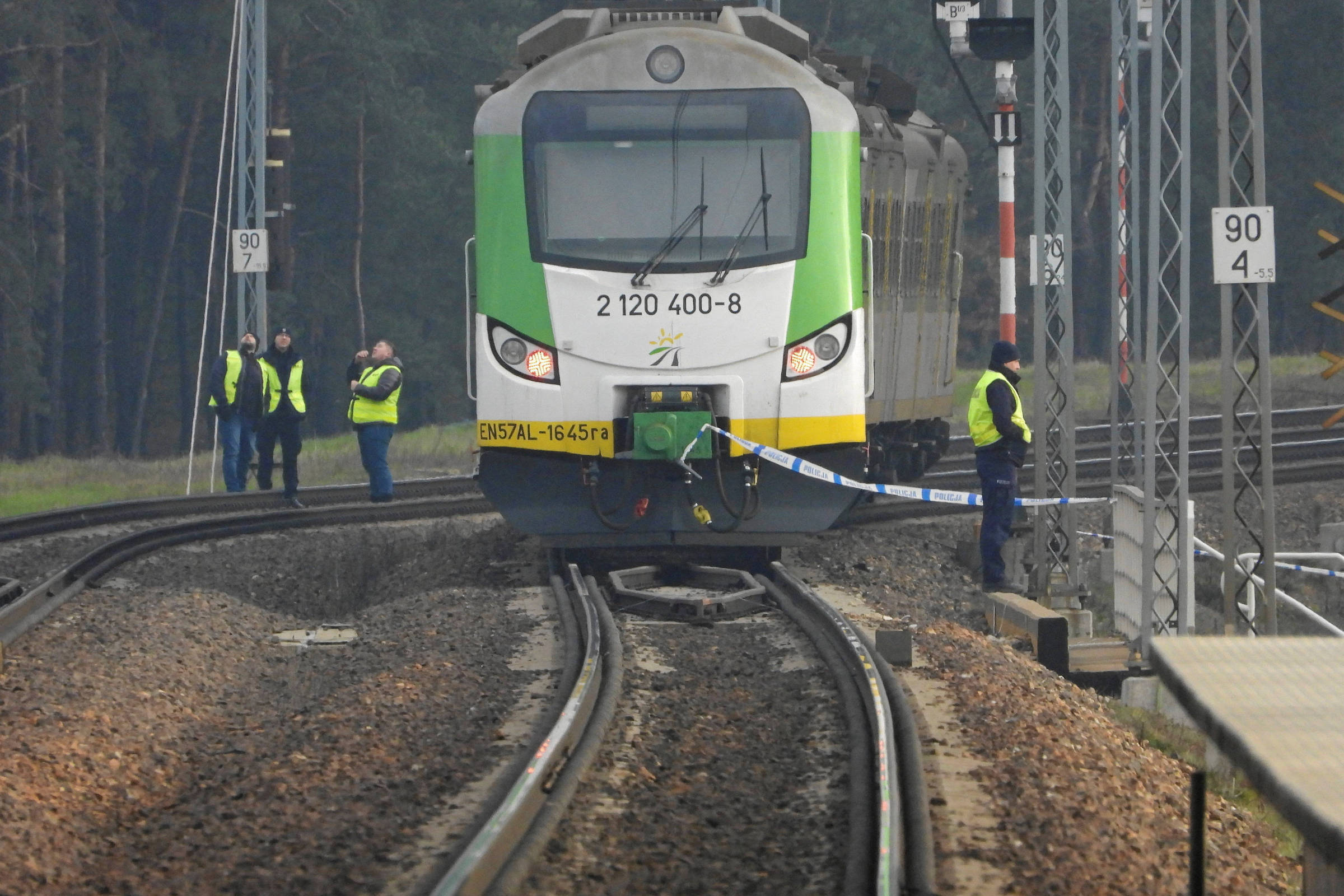 Comboio parado numa linha férrea danificada, rodeado por agentes de segurança, ilustrando a resposta das autoridades a um ato de sabotagem ferroviária na Polónia.