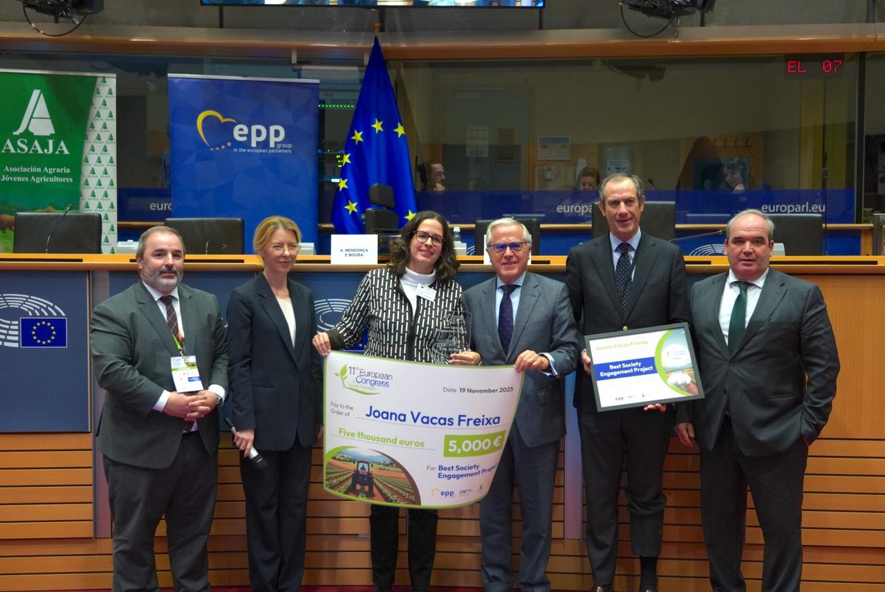 Grupo de seis pessoas em traje formal, incluindo Joana Vacas Freixa, segurando um grande diploma e um prémio, em frente a bandeiras e painéis institucionais no Parlamento Europeu, durante a cerimónia do Congresso Europeu de Jovens Agricultores.