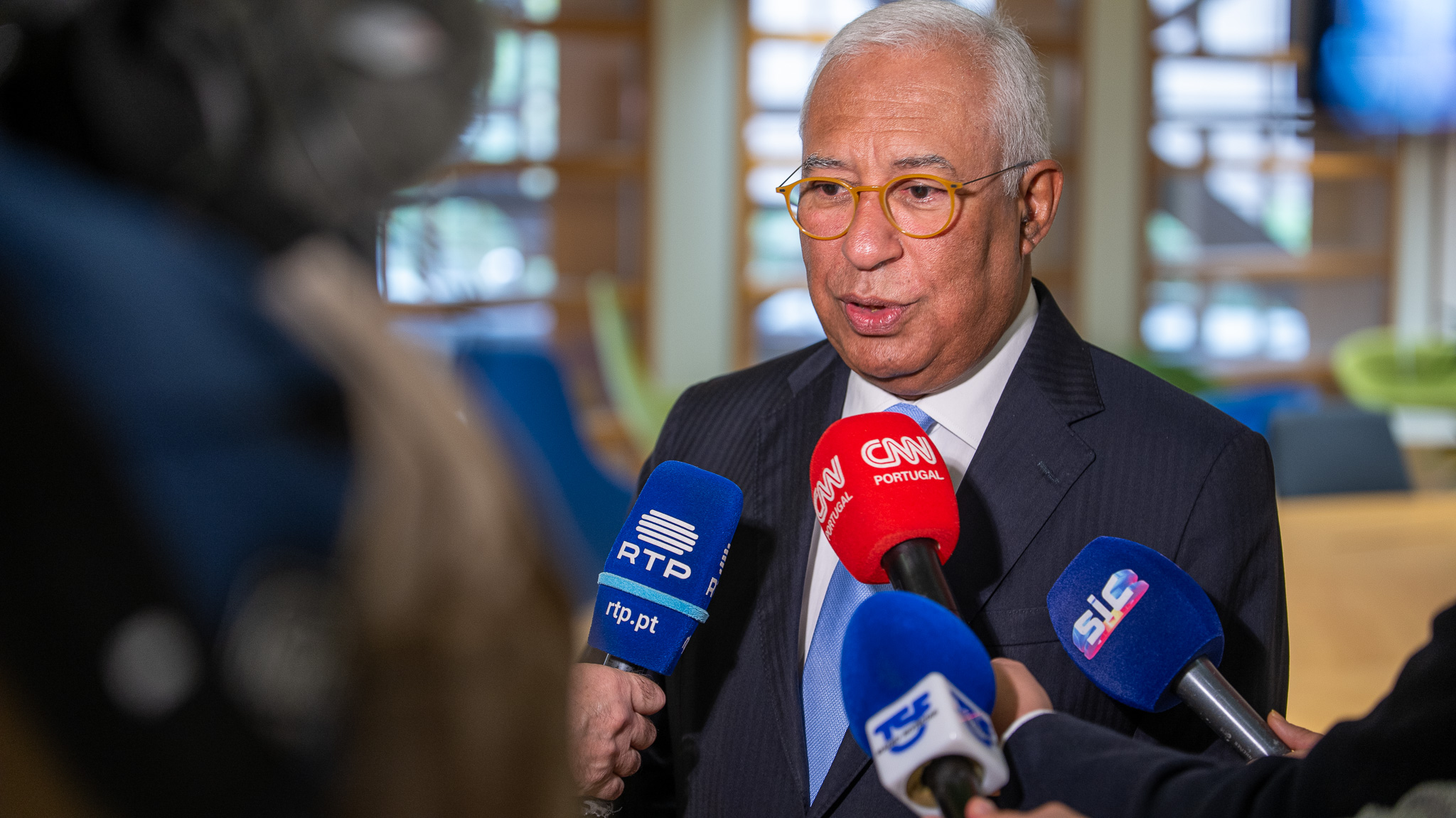 António Costa, ex-primeiro-ministro português, a dar declarações à imprensa, rodeado por microfones de vários canais de televisão.