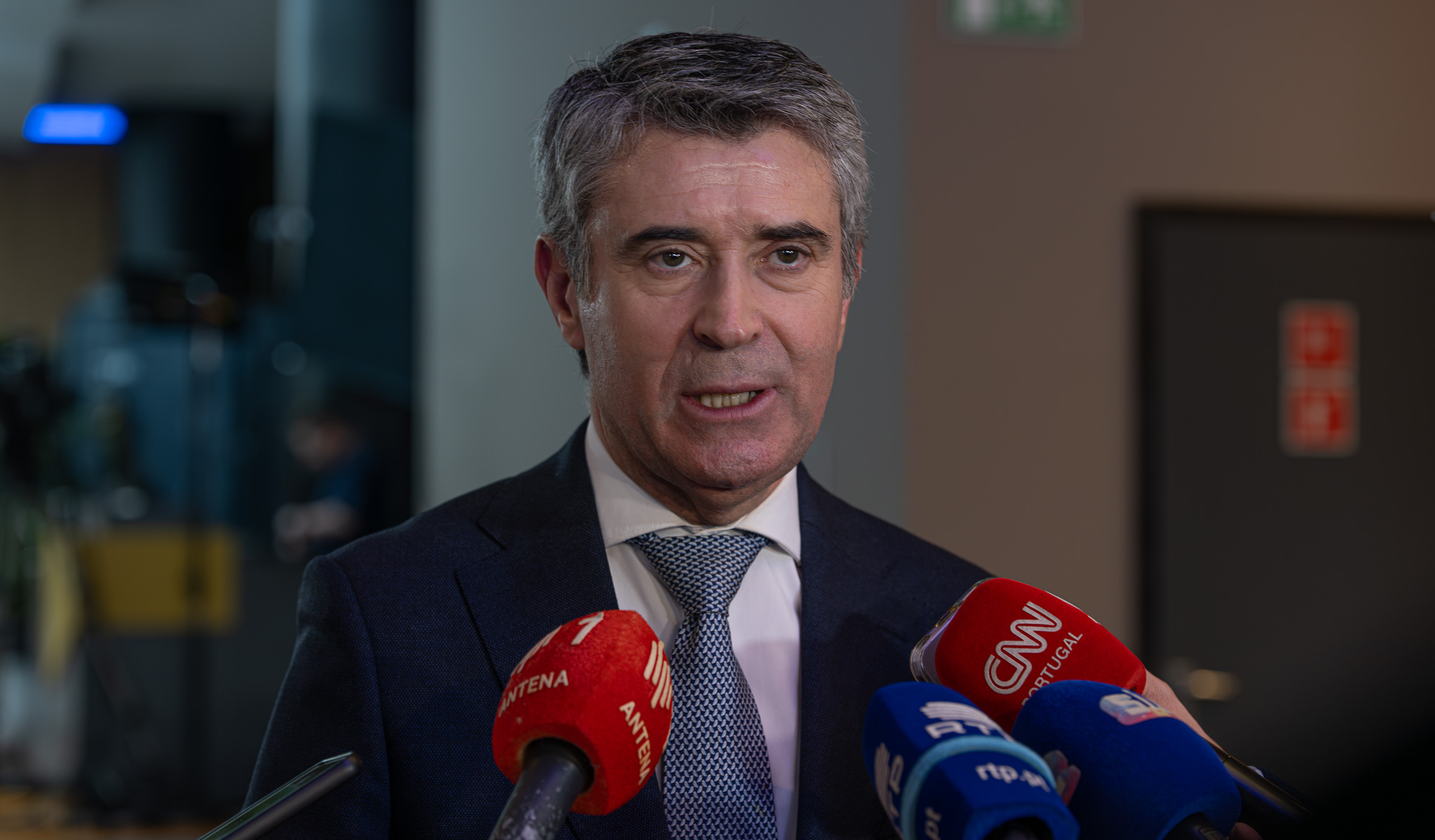 Homem de fato e gravata a falar para vários microfones de órgãos de comunicação social, durante uma declaração à imprensa.