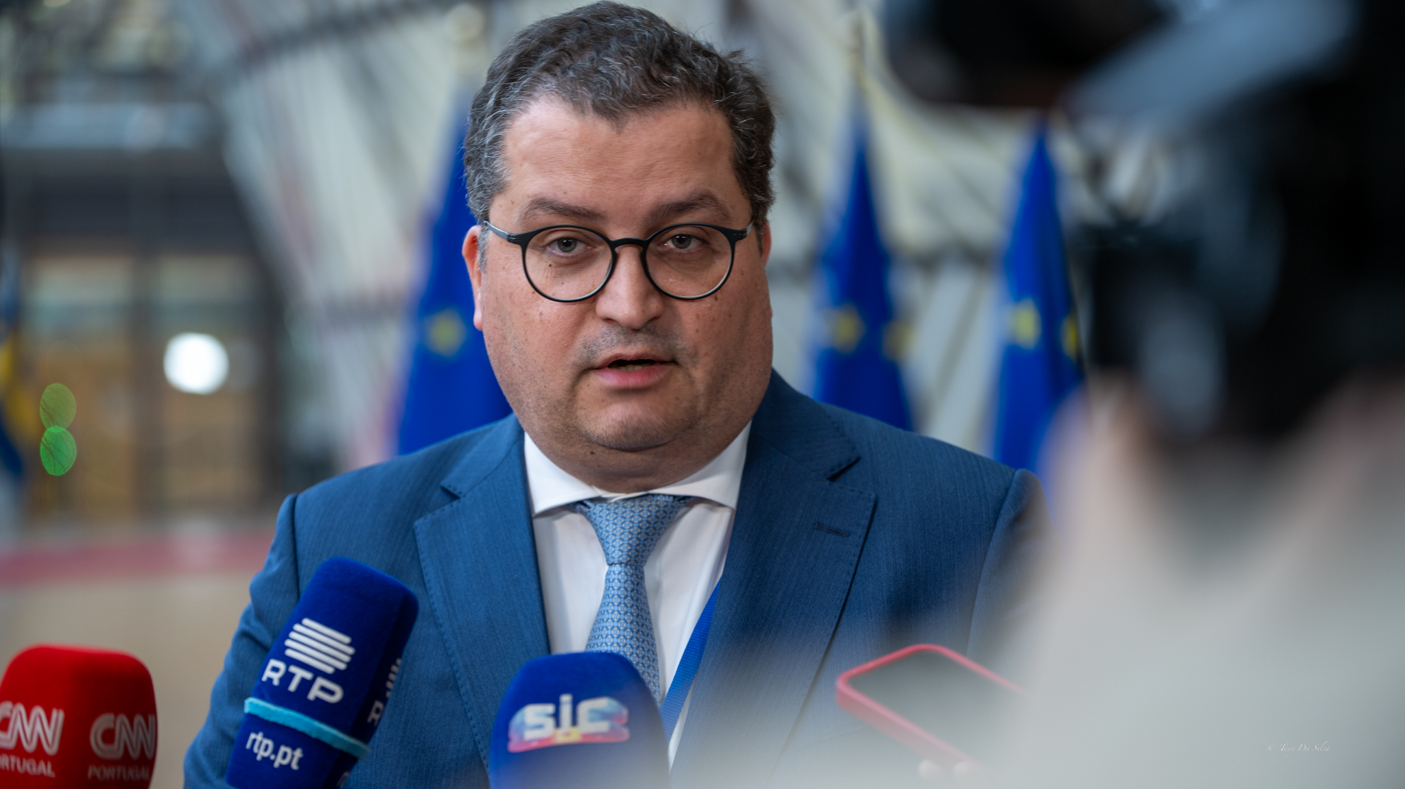 O ministro das Finanças português, Joaquim Miranda Sarmento, fala à imprensa em Bruxelas, rodeado de microfones de canais portugueses e com bandeiras da União Europeia ao fundo.