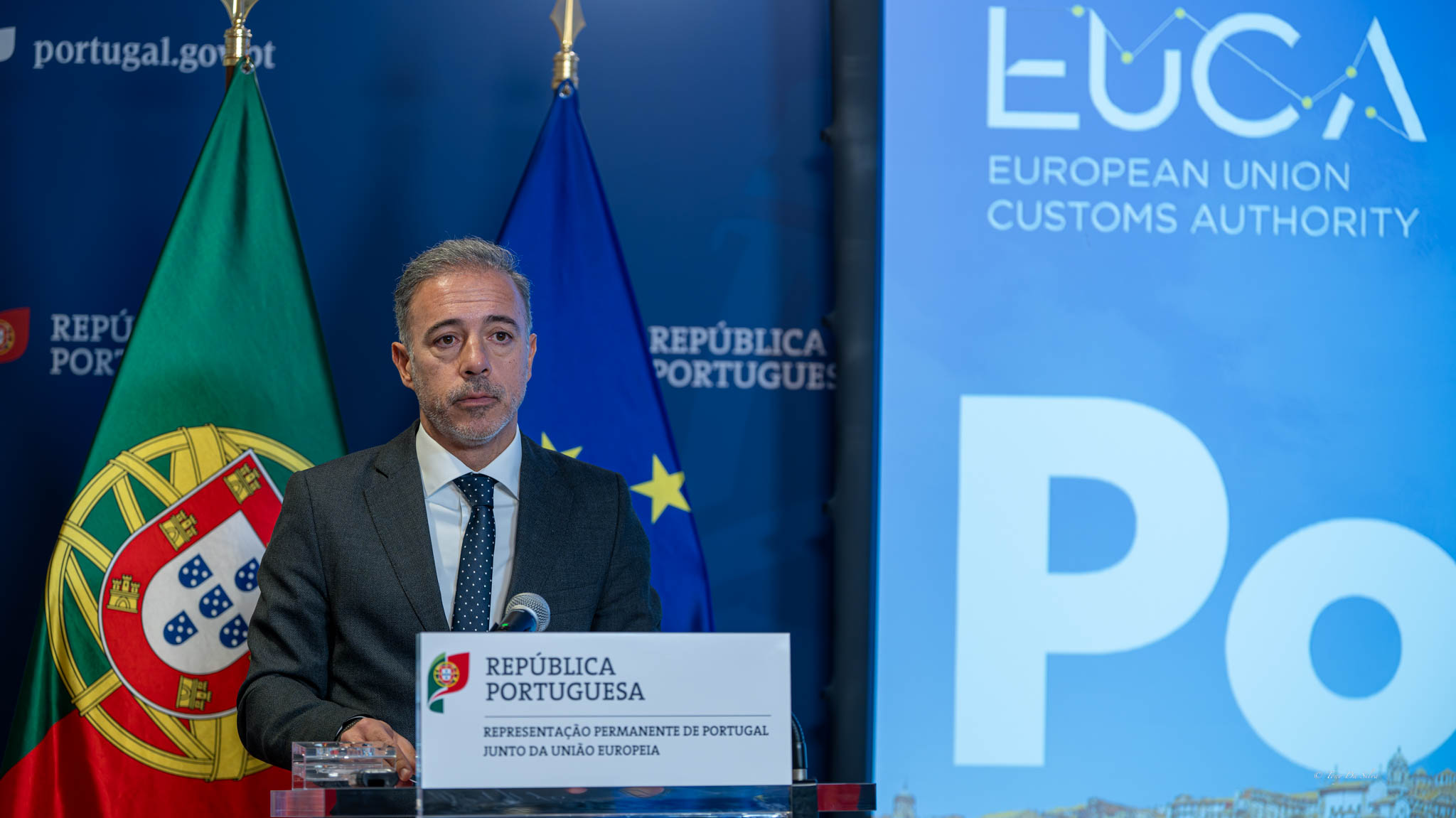 Presidente da Câmara do Porto fala num púlpito com logótipo da candidatura portuguesa à Autoridade Aduaneira Europeia, ladeado pelas bandeiras de Portugal e da União Europeia, num evento oficial em Bruxelas.