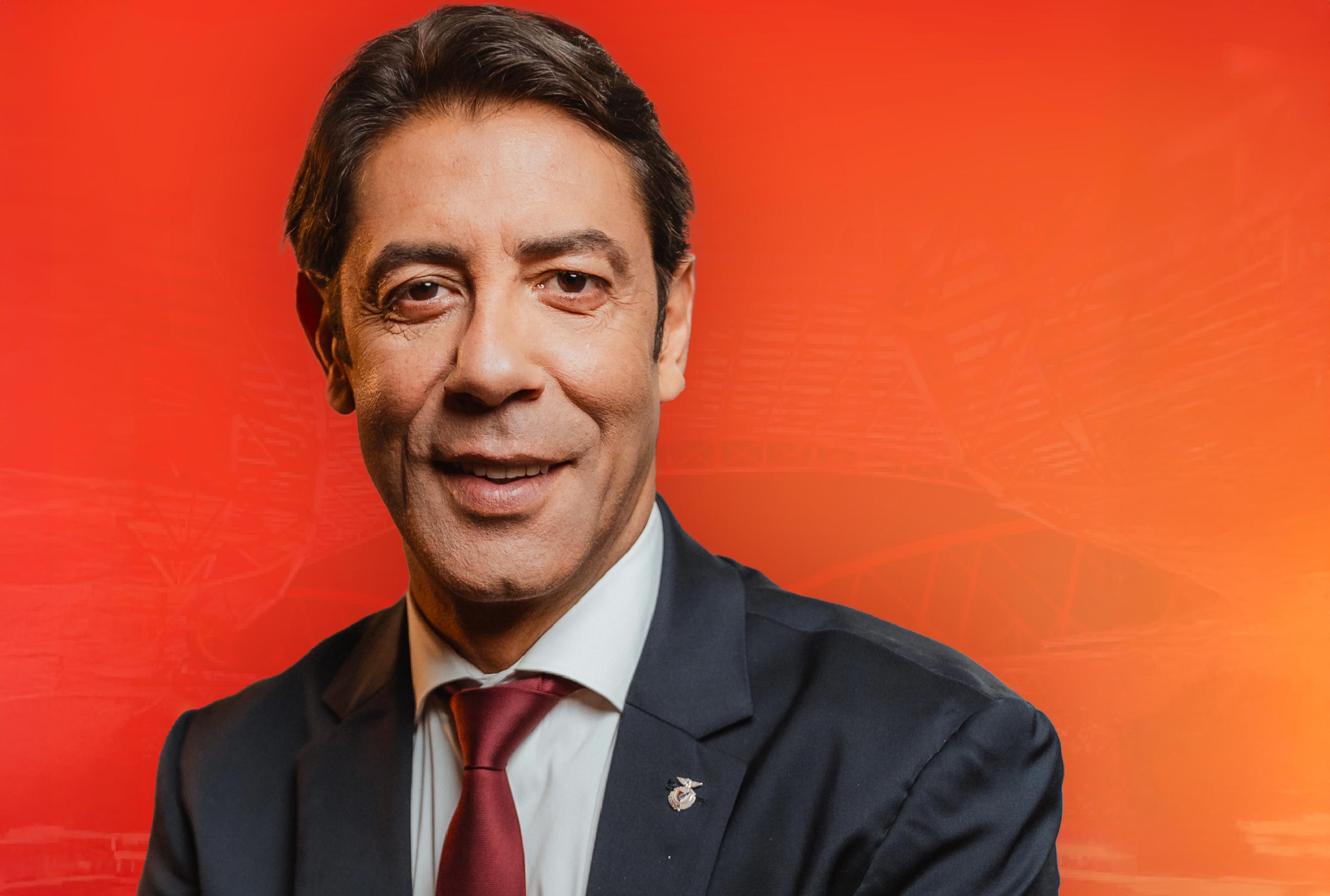Retrato formal de um homem de fato escuro e gravata vermelha, sorridente, sobre um fundo vermelho, representando liderança e ligação ao Sport Lisboa e Benfica.
