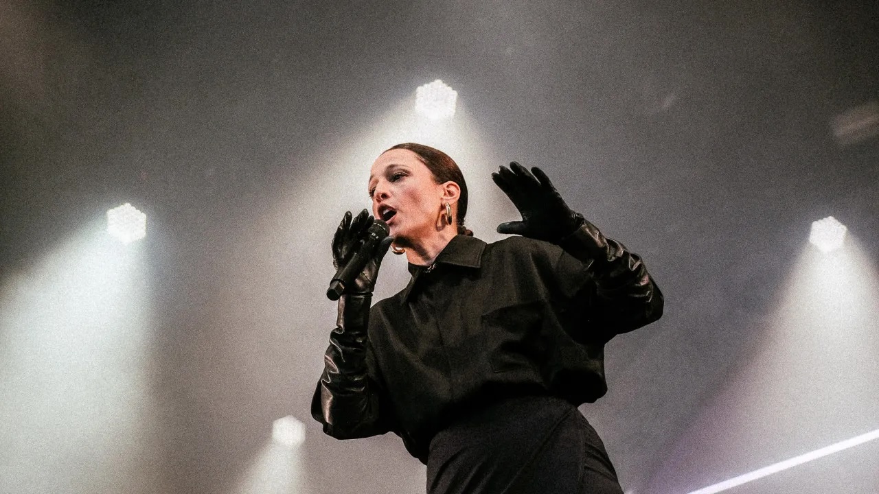 Um artista no palco, vestindo roupas pretas e luvas, cantando em um microfone com luzes brilhantes ao fundo.