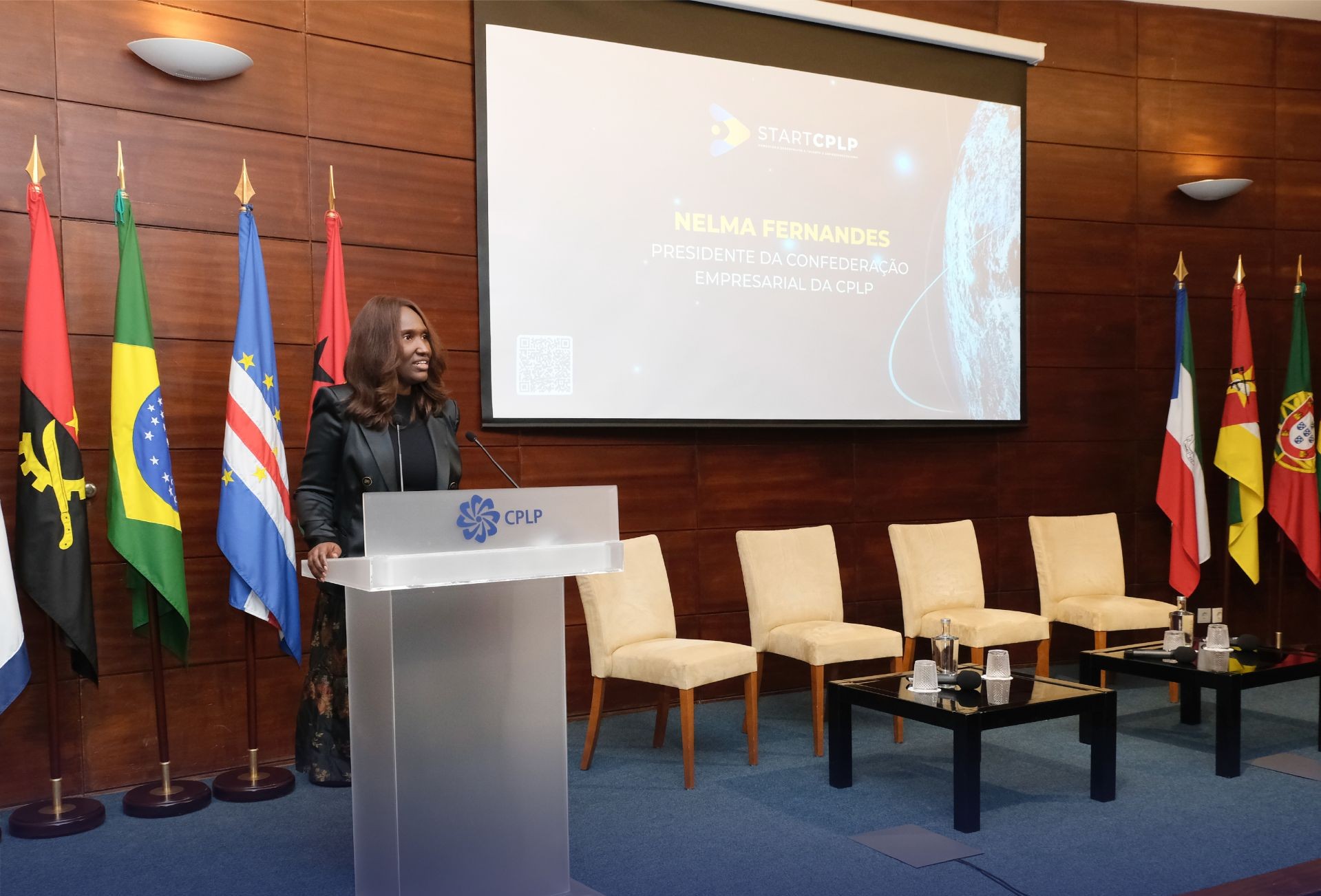 Pessoa a discursar num púlpito diante de bandeiras de países lusófonos e um ecrã de projeção, num ambiente de conferência formal.