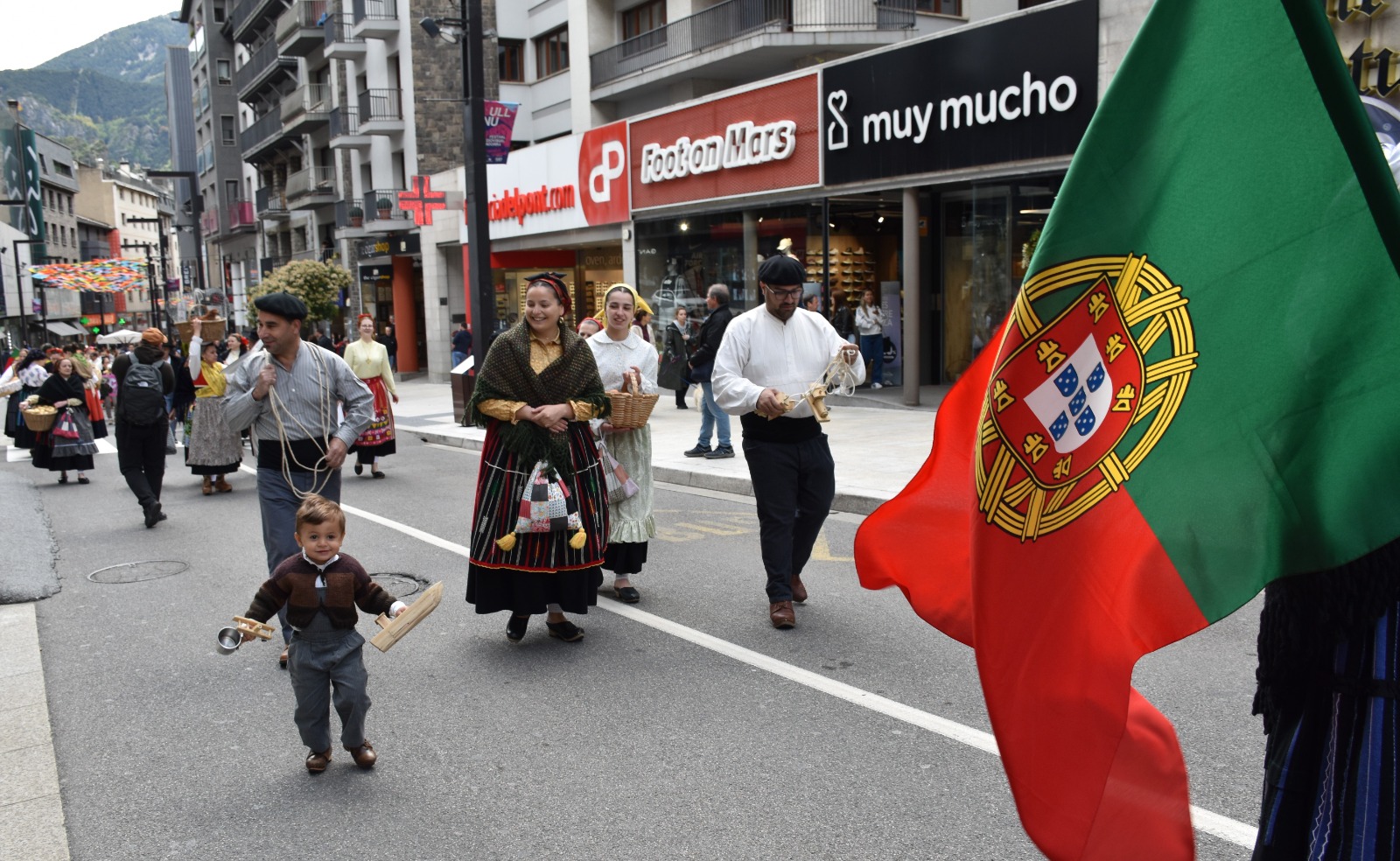 Desfile cultural em Andorra la Vella com pessoas vestidas com trajes tradicionais portugueses a caminhar na rua e uma bandeira de Portugal em destaque.