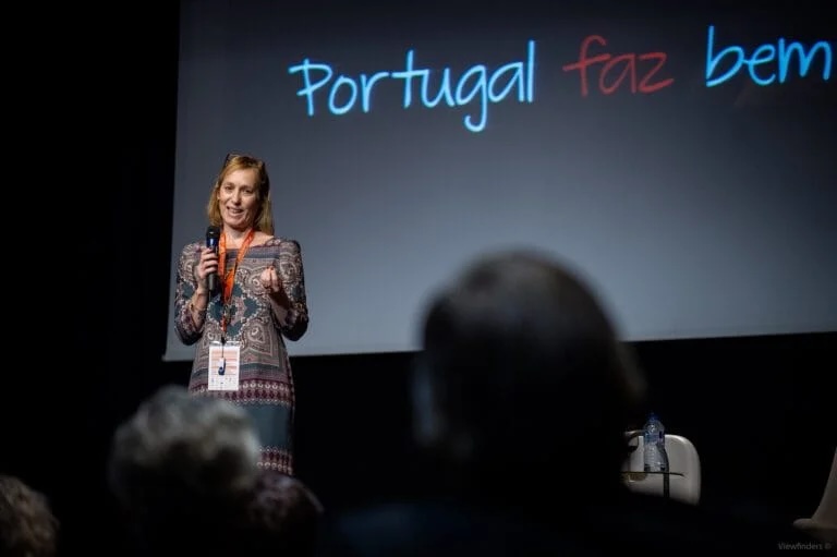 Oradora em palco durante um evento, com a frase 'Portugal faz bem' projetada no ecrã atrás, perante uma audiência.
