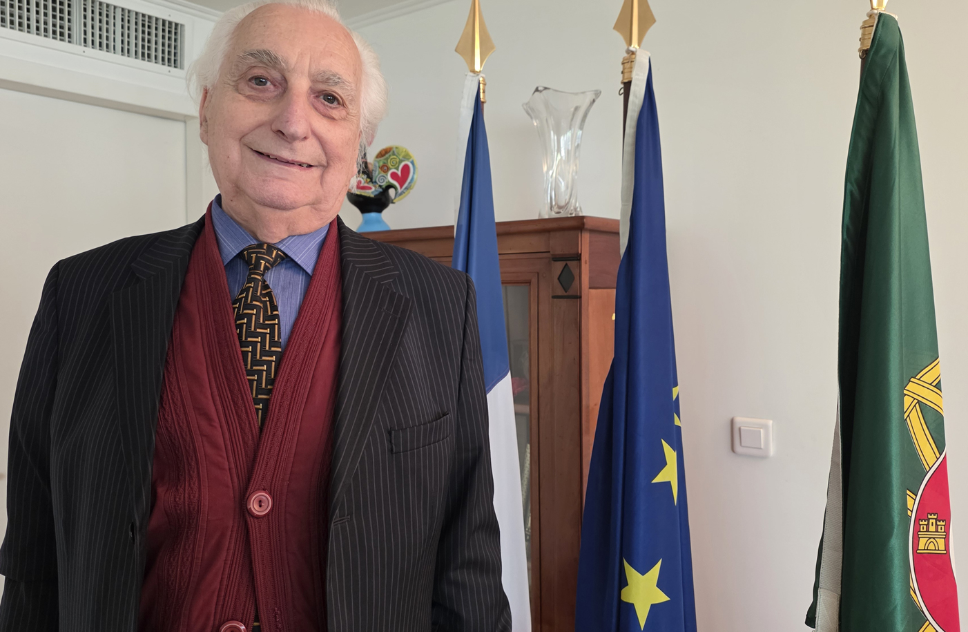 Carlos Otero, de pé e sorridente, em ambiente formal, junto a bandeiras de Portugal e da União Europeia, numa sala que sugere um espaço consular, durante a cerimónia de homenagem pelo seu contributo cultural.