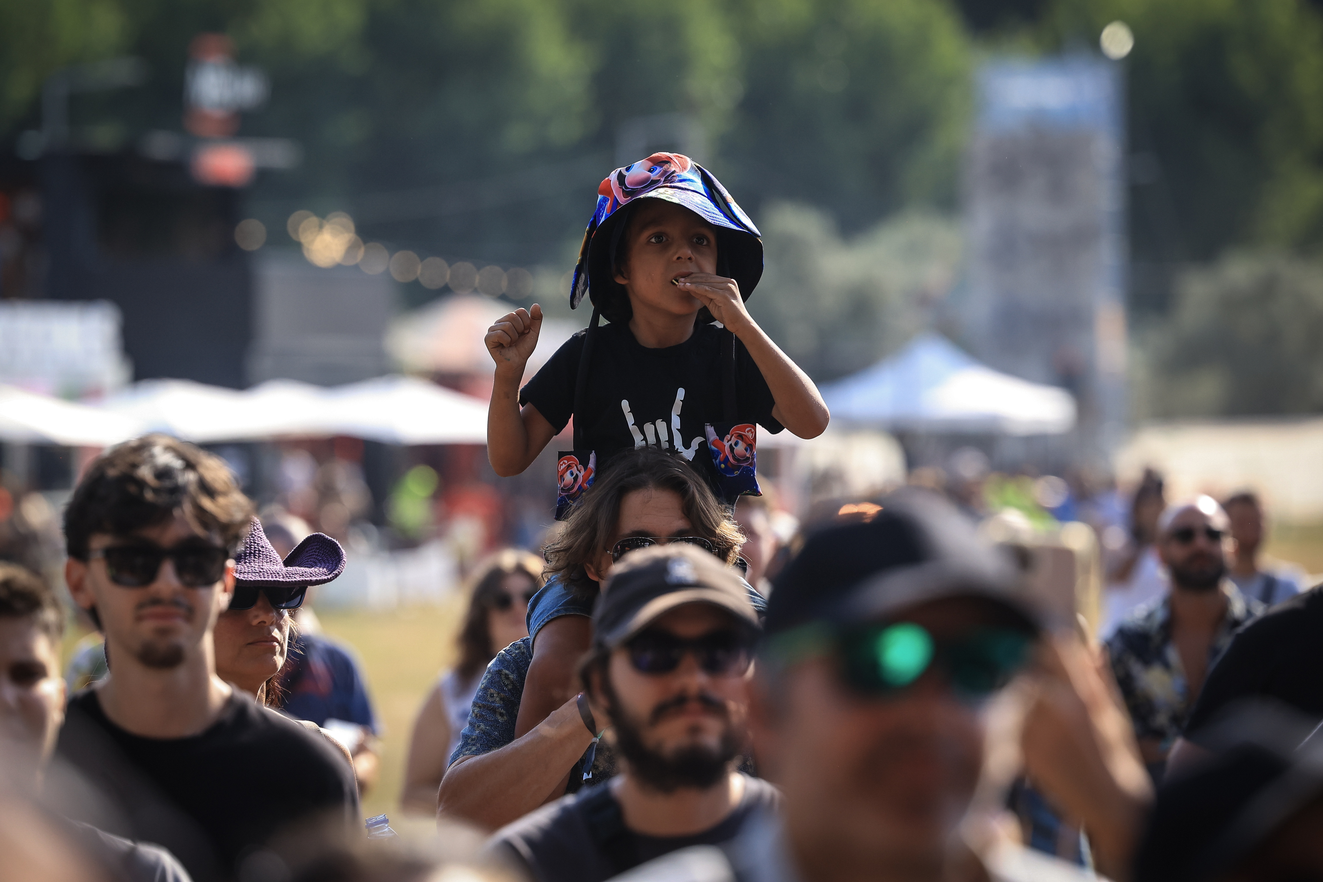 Público num festival de música ao ar livre, com várias pessoas de diferentes idades, incluindo uma criança sentada aos ombros de um adulto, num ambiente descontraído e ensolarado.