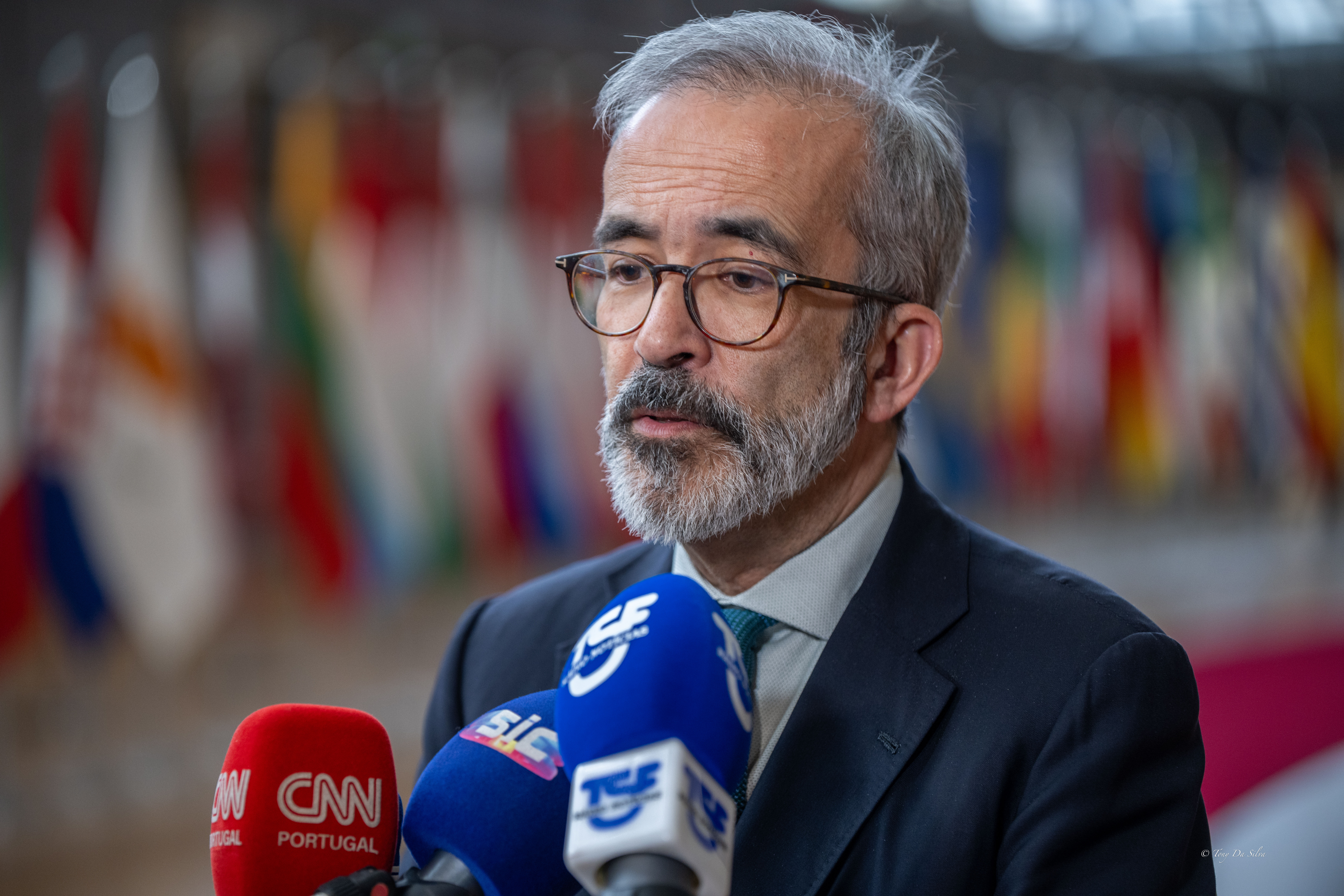 Ministro dos Negócios Estrangeiros de Portugal fala para a imprensa, com microfones à frente e bandeiras de vários países ao fundo, durante uma reunião da União Europeia em Bruxelas.