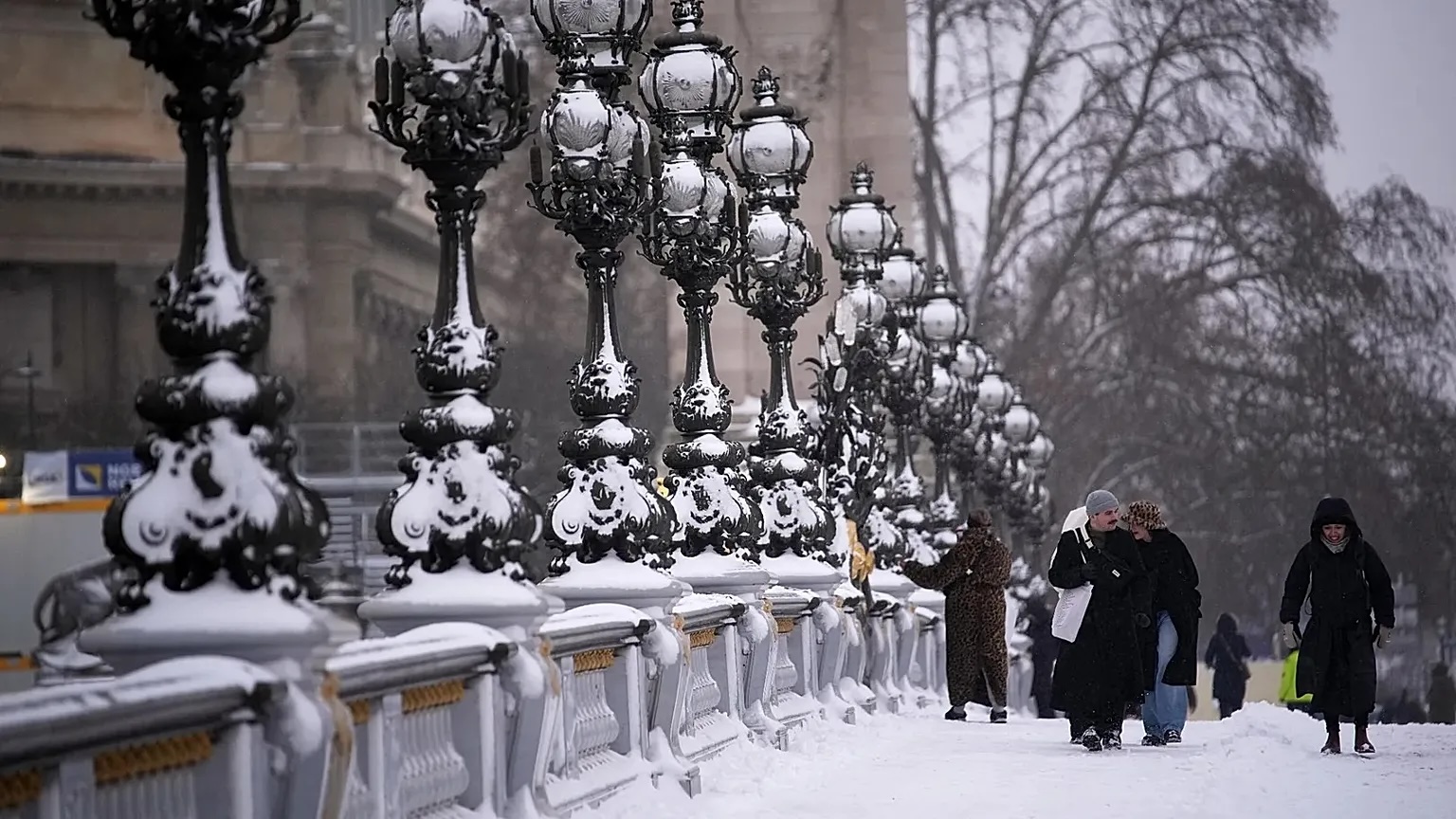 Pessoas a caminhar num passeio coberto de neve junto a uma fileira de candeeiros ornamentados.