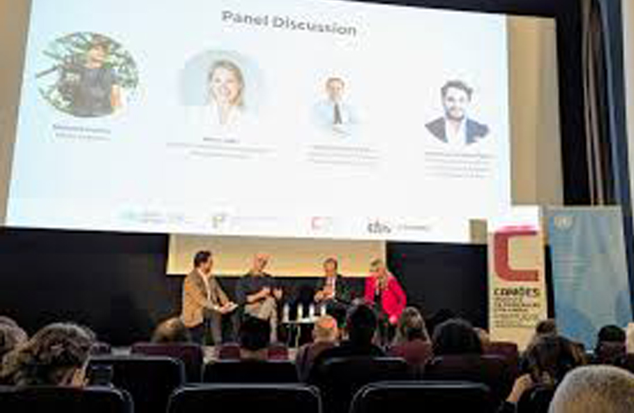 Painel de discussão com quatro pessoas sentadas em palco, moderador à esquerda, ecrã de projeção ao fundo com nomes e fotos dos oradores, público sentado a assistir. Evento celebra a língua portuguesa em Bruxelas.