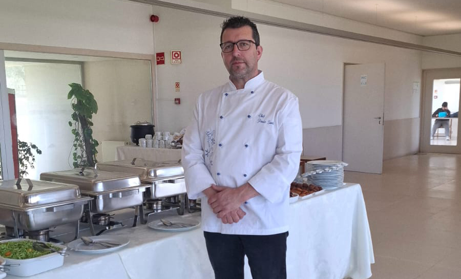 Chef com uniforme branco de cozinha junto a uma mesa de buffet com pratos e travessas, num espaço amplo e iluminado, preparado para um evento gastronómico português em Bruxelas.