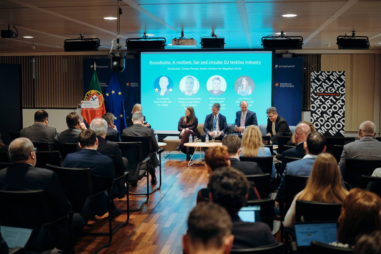 Painel de debate institucional sobre a indústria têxtil europeia, com quatro oradores sentados em palco e público atento, num auditório moderno com fundo azul e elementos visuais da União Europeia.