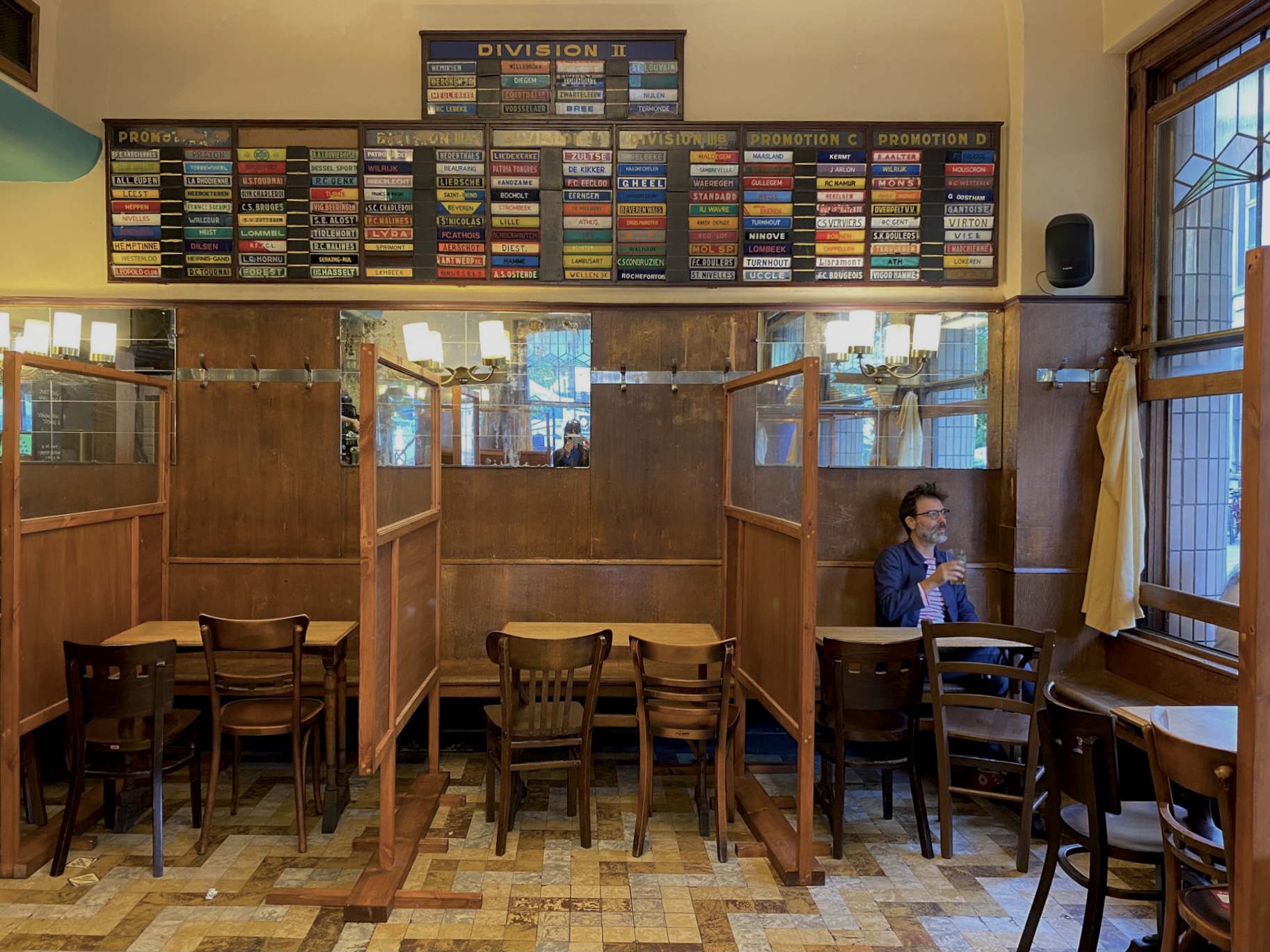 Interior de uma livraria ou café literário com mesas, cadeiras e divisórias de madeira; uma pessoa sentada lê junto à janela, e há prateleiras ou painéis coloridos na parede, sugerindo um ambiente dedicado à leitura e à literatura.