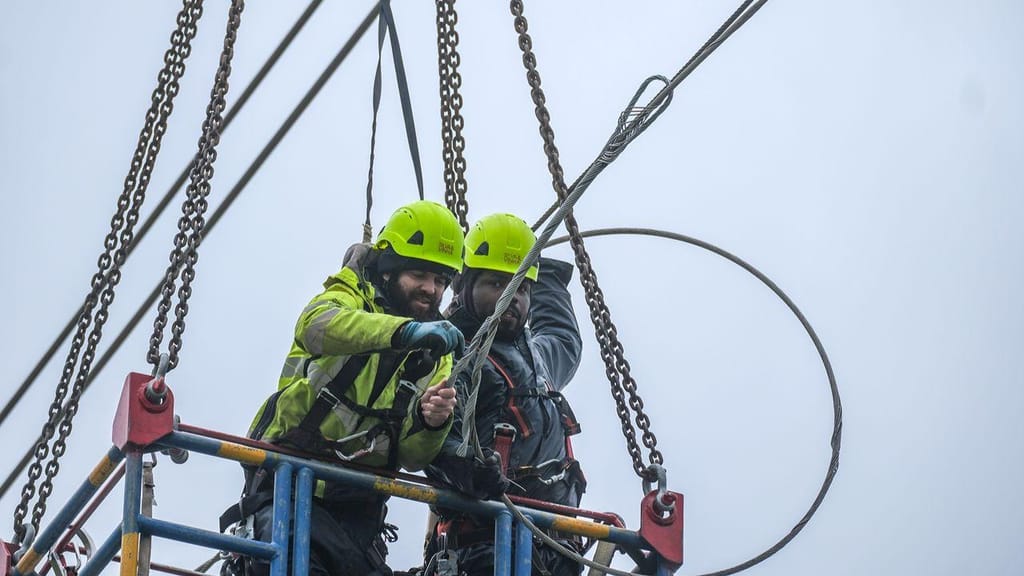 Dois trabalhadores com capacete e arnês operam numa estrutura elevada com cabos e correntes, ilustrando os esforços de restabelecimento da energia elétrica após a depressão Kristin na região de Leiria.