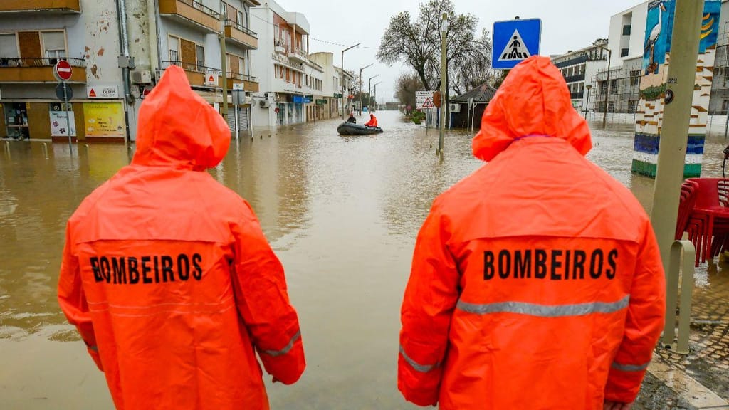 Dois bombeiros com impermeáveis laranja observam uma rua inundada, com uma equipa de resgate num barco ao fundo, ilustrando a resposta de emergência às cheias em Portugal.