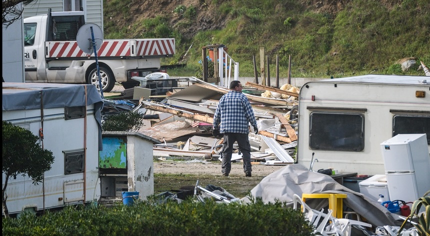 Homem de pé entre destroços de caravanas e casas móveis, avaliando danos causados por uma tempestade, com objetos espalhados e sinais de destruição visíveis.