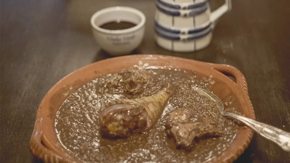 Prato de barro com arroz de cabidela, de cor escura, com pedaços de frango visíveis e uma colher, acompanhado por uma chávena e um jarro ao fundo, evocando uma refeição tradicional portuguesa.